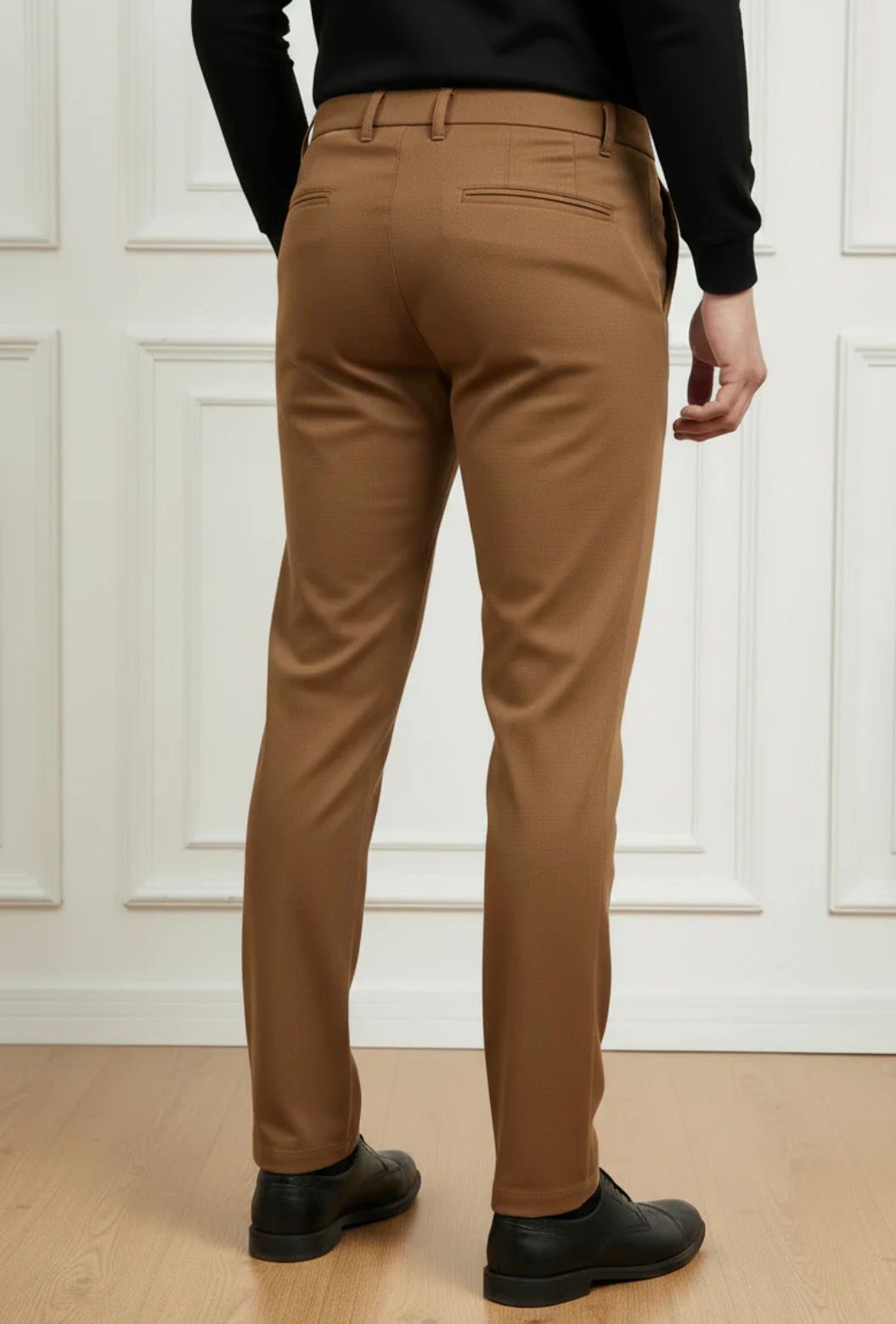Person wearing brown pants and black shoes against a white paneled wall.